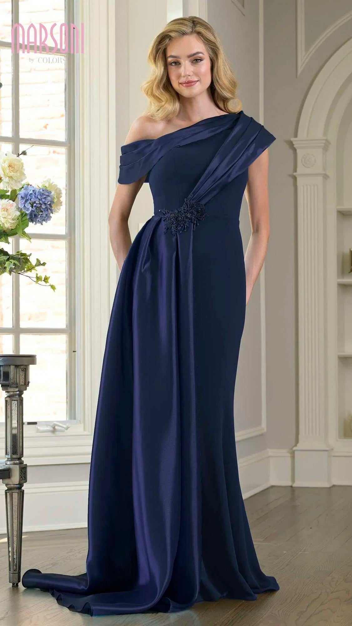 A woman stands indoors wearing the Marsoni M346, a navy long formal dress featuring a floor-length, one-shoulder stretch crepe design with floral embellishment at the waist.